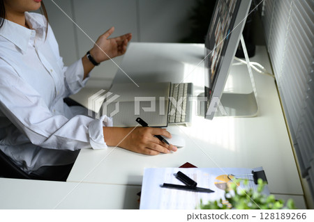 Businesswoman participating in an online video call, taking notes at modern workstation Businesswoman participating in an online video call, taking notes at modern workstation 128189266