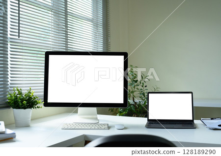 Modern home office with desktop monitor and laptop, both displaying blank white screens 128189290
