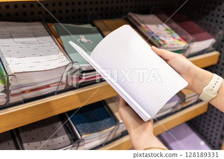 Customer hands hold an open blank grid notebook, choosing new school or office stationery supplies from a well-stocked retail store shelf 128189331