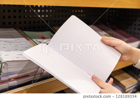 Person's hands carefully holding an open blank grid-lined notebook in a stationery store, ideal for students, office supplies, or creative planning Person's hands carefully holding an open blank grid-lined notebook in a stationery store, ideal for students, office supplies, or creative planning 128189334