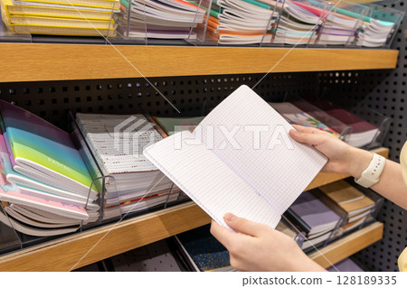 Buyer hands holding an open grid-lined notebook, exploring diverse stationery items on retail shelves for academic or office supply needs 128189335