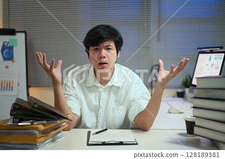 Tired businessman expressing stress and confusion during overtime hours in a cluttered office 128189381