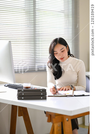 Asian woman analyzing data and writing on paper in a bright modern office Asian woman analyzing data and writing on paper in a bright modern office 128189419