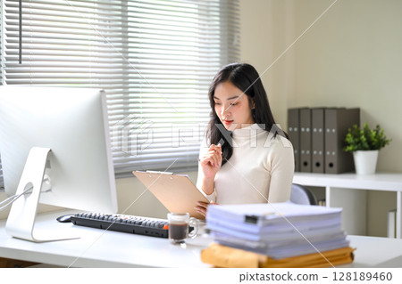 Smiling Asian businesswoman working at desk, reviewing documents during a video call Smiling Asian businesswoman working at desk, reviewing documents during a video call 128189460