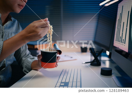 Close up of man eating instant noodles at desk while working late at night under office lighting 128189463