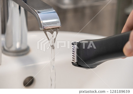 A hand cleaning an electric trimmer head under a flowing chrome faucet in a bathroom sink, promoting daily personal hygiene and careful grooming 128189639