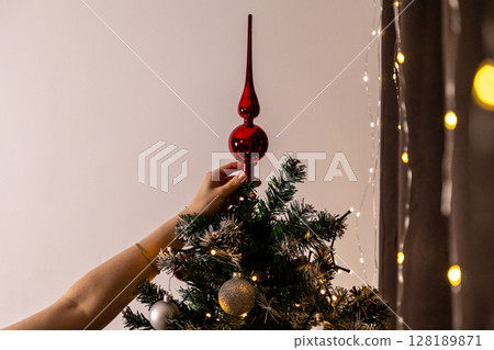 placing a shiny red christmas tree topper on a decorated artificial tree with warm string lights and festive ornaments in a cozy interior 128189871