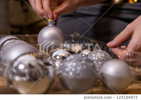 placing a matte silver christmas ornament into a plastic tray filled with shiny and glittery baubles, close-up with soft warm lighting and blurred festive background placing a matte silver christmas ornament into a plastic tray filled with shiny and glittery baubles, close-up with soft warm lighting and blurred festive background 128189872