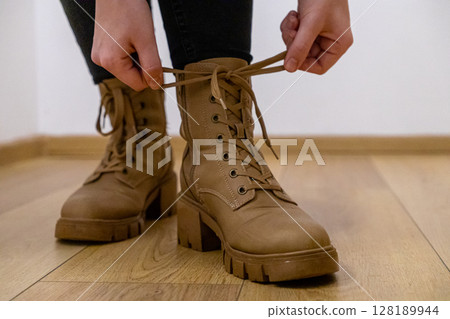 tying laces on stylish brown leather boots with chunky heels indoors, close-up of hands and detailed view of footwear on wooden floor tying laces on stylish brown leather boots with chunky heels indoors, close-up of hands and detailed view of footwear on wooden floor 128189944