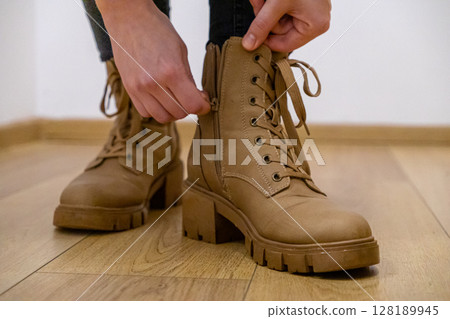 zipping up brown leather boots with laces and chunky heels indoors, close-up of hands adjusting the zipper on the footwear over a wooden floor 128189945