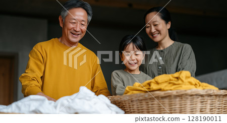 Asian family folding laundry together in sunlit room, showing teamwork, shared responsibility and daily life happiness 128190011
