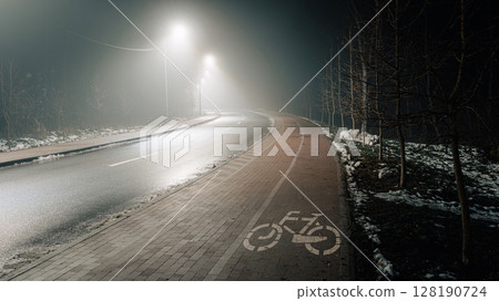 Empty road and bike path in a foggy nighttime setting, illuminated by streetlights. 128190724