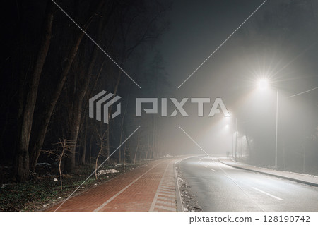 Foggy urban road and red-brick bike lane lit by soft streetlights, surrounded by shadowy trees. Foggy urban road and red-brick bike lane lit by soft streetlights, surrounded by shadowy trees. 128190742
