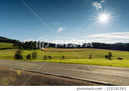Foothills of the Austrian Alps with asphalt road on foreground and sun on blue morning sky Foothills of the Austrian Alps with asphalt road on foreground and sun on blue morning sky 128191263