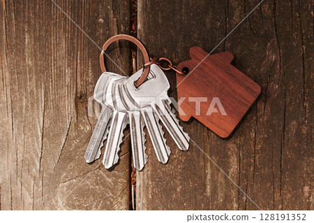 Metal home keys with wooden house-shaped keychain on weathered garden table. Close-up shot 128191352