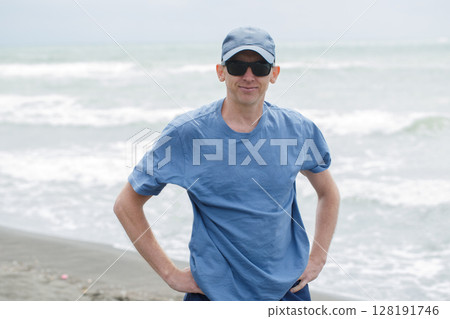 Man Wearing Sunglasses and a Blue Shirt Standing on the Beach Enjoying the Sea View 128191746