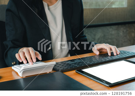 A business professional working with a calculator and keyboard in a modern office environment, featuring a tablet. 128191756