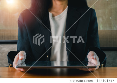 A business professional in a suit holding a tablet, sitting at a wooden desk with sunlight streaming in. 128191757