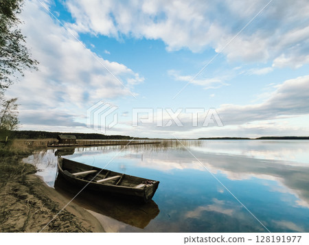 Old wooden fishing boat moored on shore of calm lake reflecting cloudy sky during beautiful sunny day, with small wooden pier and hut on background. Old wooden fishing boat anchored beside serene lake 128191977