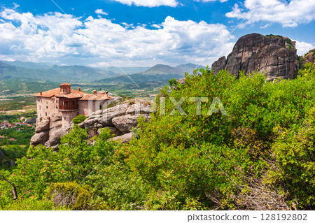 Ancient Meteora monastery perched on cliff edge beside massive rock formation in Greece 128192802