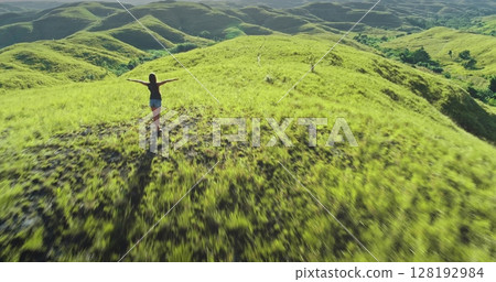 Aerial view of a young female tourist embracing the beauty of nature in Sumba Island, Indonesia, with her arms outstretched against the backdrop of rolling green hills Aerial view of a young female tourist embracing the beauty of nature in Sumba Island, Indonesia, with her arms outstretched against the backdrop of rolling green hills 128192984