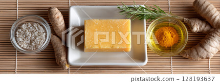 A natural turmeric soap bar surrounded by herbs, spices, and salt on a bamboo mat, showcasing organic skincare ingredients and textures 128193637