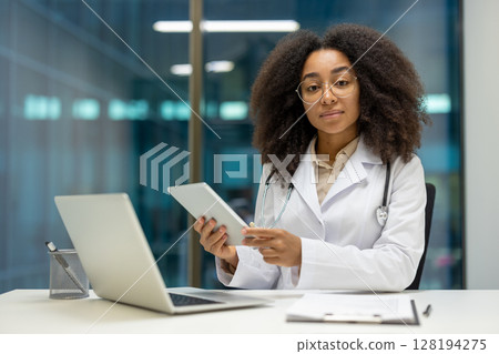 Portrait of serious confident female doctor at workplace inside modern clinic office. Female doctor looking at camera with tablet computer in hands sitting at desk. Portrait of serious confident female doctor at workplace inside modern clinic office. Female doctor looking at camera with tablet computer in hands sitting at desk. 128194275
