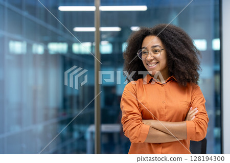 Young adult African American woman stands confidently with crossed arms in a modern office setting, smiling and looking away 128194300