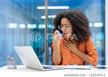 An office worker is experiencing an asthma attack. She struggles to breathe while reaching for her inhaler near her open laptop and notebook. 128194311