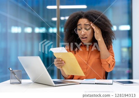 Sad unhappy woman holding mail envelope message in hands. Office worker reading notification letter with bad news, sitting at desk inside office. 128194313