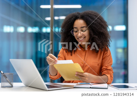 A cheerful young woman sits at her desk, holding a paper and smiling. She looks happy while examining the contents of the envelope. A cheerful young woman sits at her desk, holding a paper and smiling. She looks happy while examining the contents of the envelope. 128194314