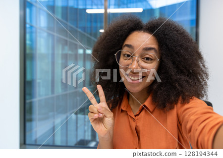 A happy young woman with afro hair and round glasses shows the peace sign during a selfie video call in a modern office 128194344