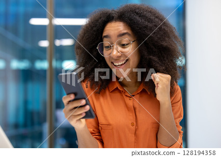 An excited young woman in glasses celebrates while looking at her phone, expressing joy and success in a modern office setting. An excited young woman in glasses celebrates while looking at her phone, expressing joy and success in a modern office setting. 128194358
