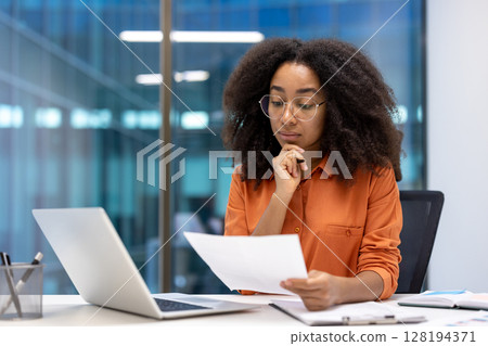 Serious thinking woman financier doing paperwork inside office. Focused accountant holding documents and papers in hands. Businesswoman checking data, working inside office. 128194371