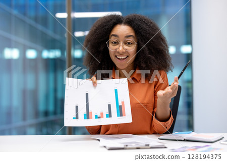 A smiling woman shows a graph, possibly during a presentation or meeting, in a modern office setting. She is wearing glasses. A smiling woman shows a graph, possibly during a presentation or meeting, in a modern office setting. She is wearing glasses. 128194375