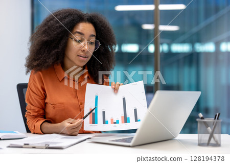 A focused woman presents a data chart to an audience via laptop during a video conference in a modern office setting. She holds a pen for emphasis. 128194378