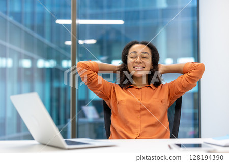 A relaxed businesswoman takes a moment to unwind in her office with hands behind her head, smiling. Focus on her serenity. A relaxed businesswoman takes a moment to unwind in her office with hands behind her head, smiling. Focus on her serenity. 128194390