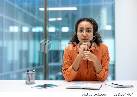 A young woman with a headset sits at a desk in an office setting, looking directly at the camera. She is ready to assist clients. 128194404