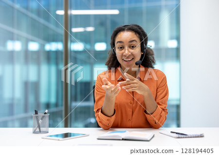 A young woman wearing a headset gestures while engaging in a video call, working in a modern office environment. Her bright smile conveys friendliness. 128194405