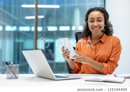 A smiling African American woman wearing a headset and glasses uses a tablet at her desk, ready to assist customers 128194409