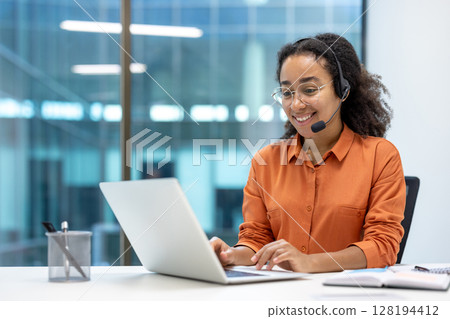 Smiling customer service representative wearing a headset and glasses working on a laptop in a modern office setting. Focus on communication and support. Smiling customer service representative wearing a headset and glasses working on a laptop in a modern office setting. Focus on communication and support. 128194412