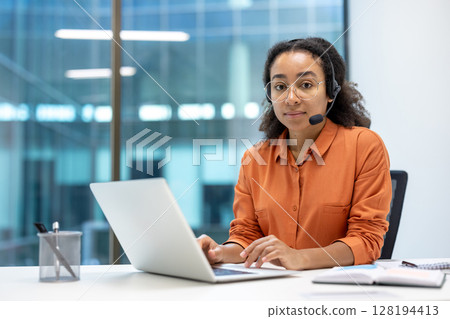 Portrait of serious focused woman with headset, customer support worker inside office at workplace, using laptop for video call. Looking thoughtfully at camera. Portrait of serious focused woman with headset, customer support worker inside office at workplace, using laptop for video call. Looking thoughtfully at camera. 128194413