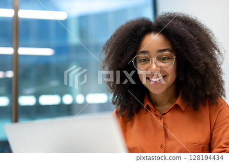 A beautiful young woman with curly hair and glasses smiles at the camera while working on a laptop in an office setting. A beautiful young woman with curly hair and glasses smiles at the camera while working on a laptop in an office setting. 128194434