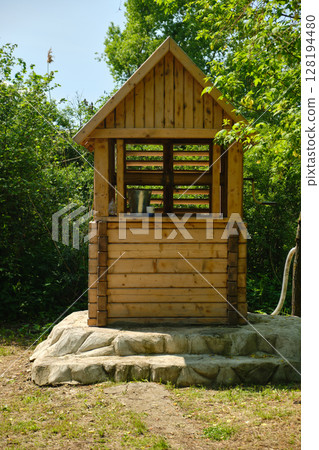 rustic wooden water well with a bucket inside stands on stone steps surrounded by vibrant green 128194480