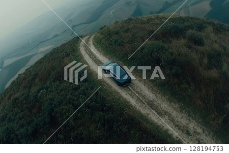Aerial view of a dark car driving on a winding dirt road through green hills, showcasing remote travel and adventure in a misty mountainous landscape Aerial view of a dark car driving on a winding dirt road through green hills, showcasing remote travel and adventure in a misty mountainous landscape 128194753
