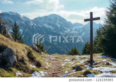 Wooden cross on a snowy mountain trail with evergreen trees and snow-capped peaks in the background under a bright sky 128194965