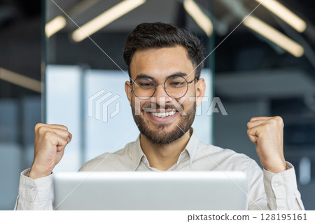 A young, cheerful man with glasses celebrates a win in front of a laptop, clenched fists in the air, in an office setting. A young, cheerful man with glasses celebrates a win in front of a laptop, clenched fists in the air, in an office setting. 128195161