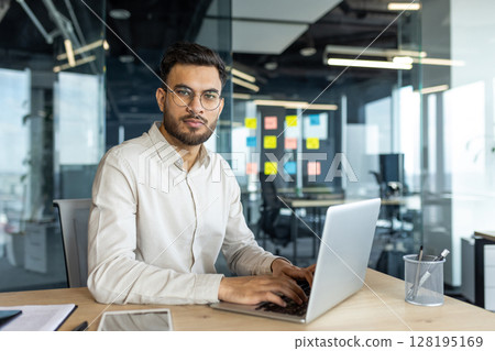 Portrait of mature and serious businessman at workplace. Man in shirt looking at camera, working with laptop inside office. Portrait of mature and serious businessman at workplace. Man in shirt looking at camera, working with laptop inside office. 128195169