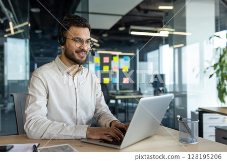A smiling man with glasses and a headset works on a laptop at an office desk. He appears to be engaged in a video call. A smiling man with glasses and a headset works on a laptop at an office desk. He appears to be engaged in a video call. 128195206