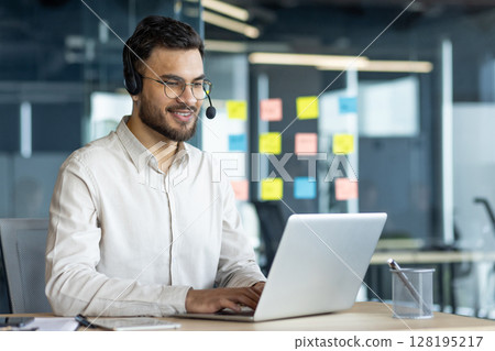 Latin American businessman working efficiently on a laptop wearing a headset in office environment, demonstrating effective remote communication, work engagement, and business professionalism. 128195217